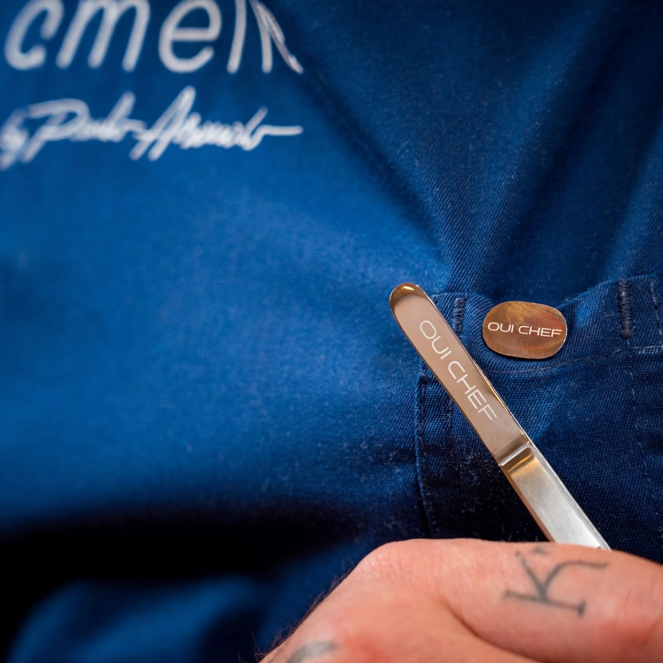 Chef wearing a blue apron from Amelia restaurant holding kitchen tweezers to the magnetic clip pin engraved with 'Oui Chef'
