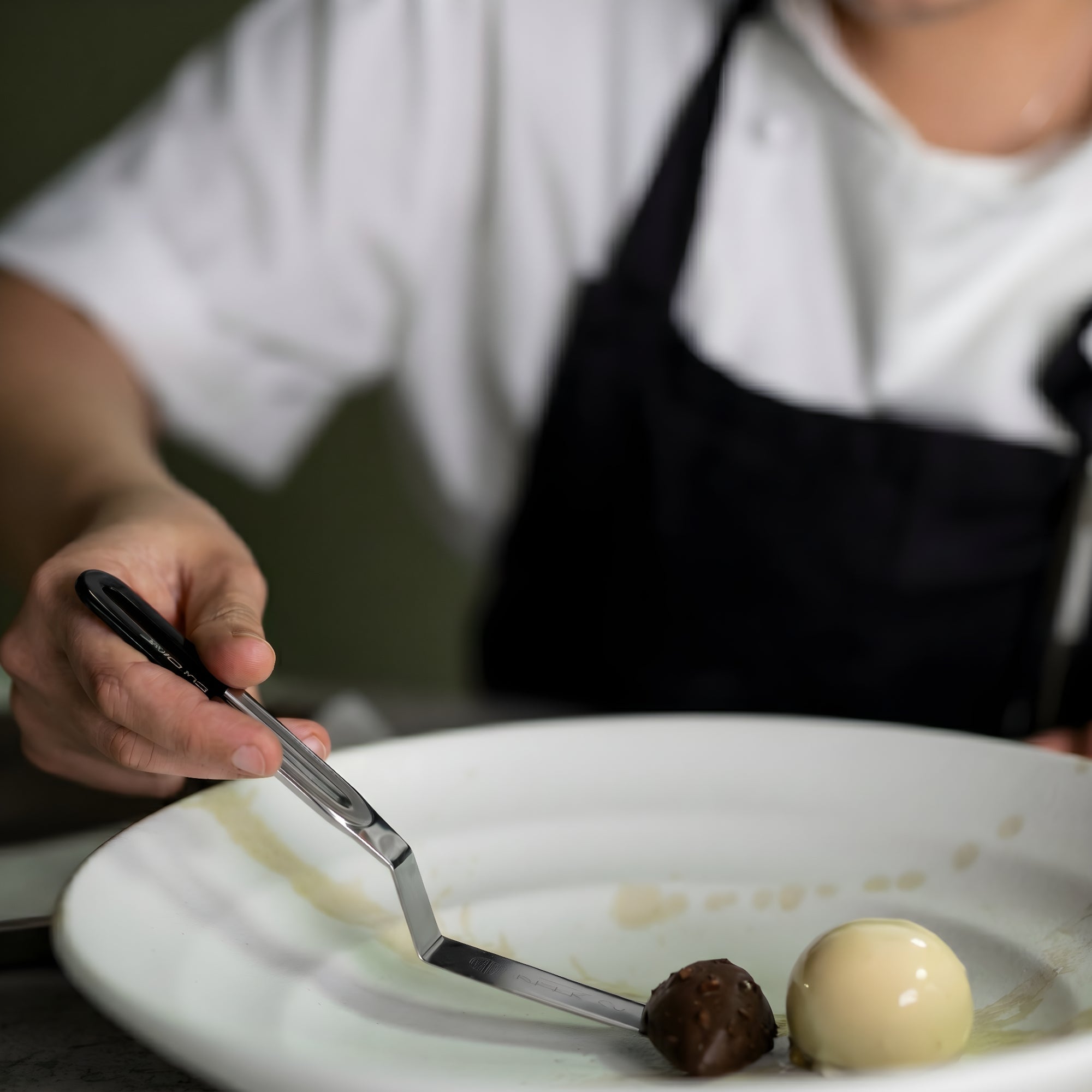 Chef delicately plating a piece of artisanal chocolate beside a quenelle of ice cream using a palette knife.