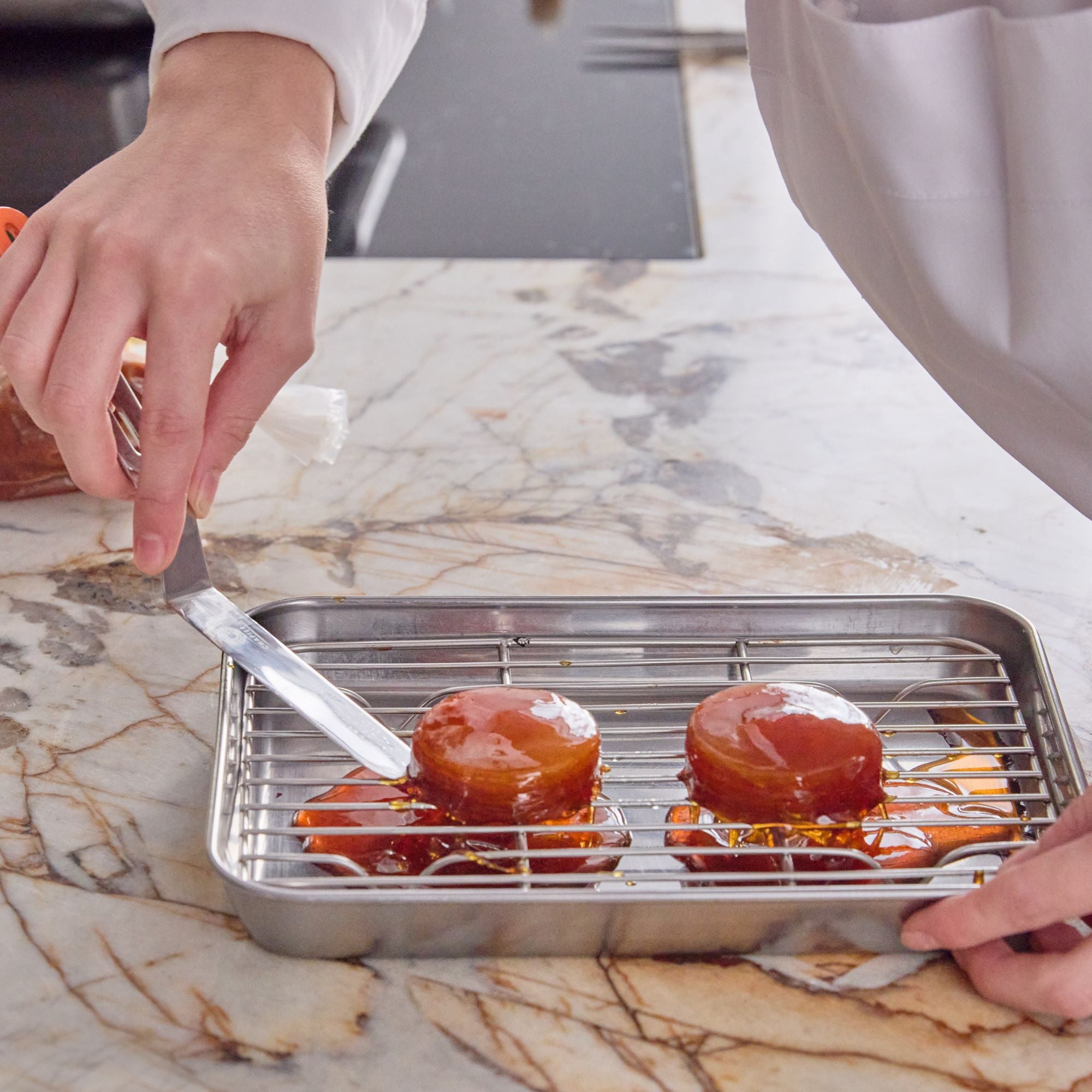 A Chef holding a stainless steel palette knife and carefully gliding under a delicate dessert that's sitting on a tray on a marble kitchen countertop.
