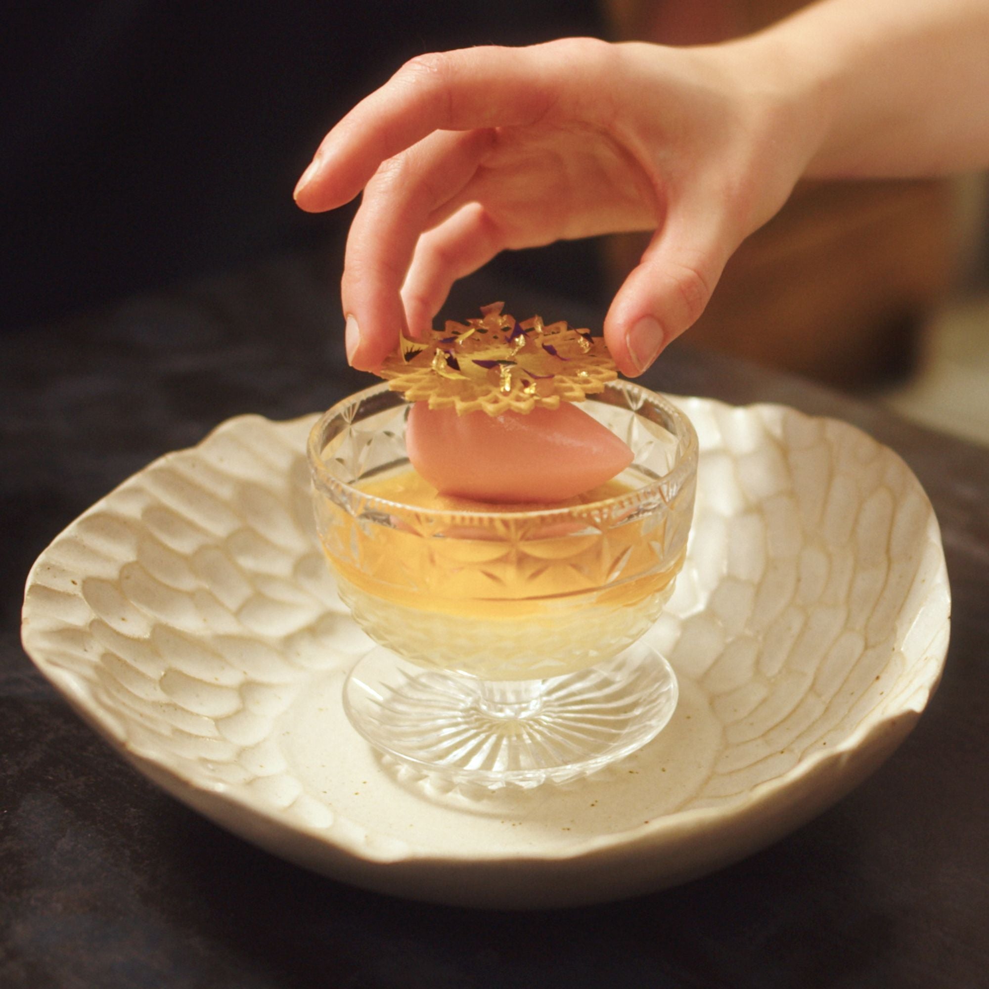 Hand placing a decorative finish on a pink rocher of ice cream presented in a glass container, on a textured white plate.