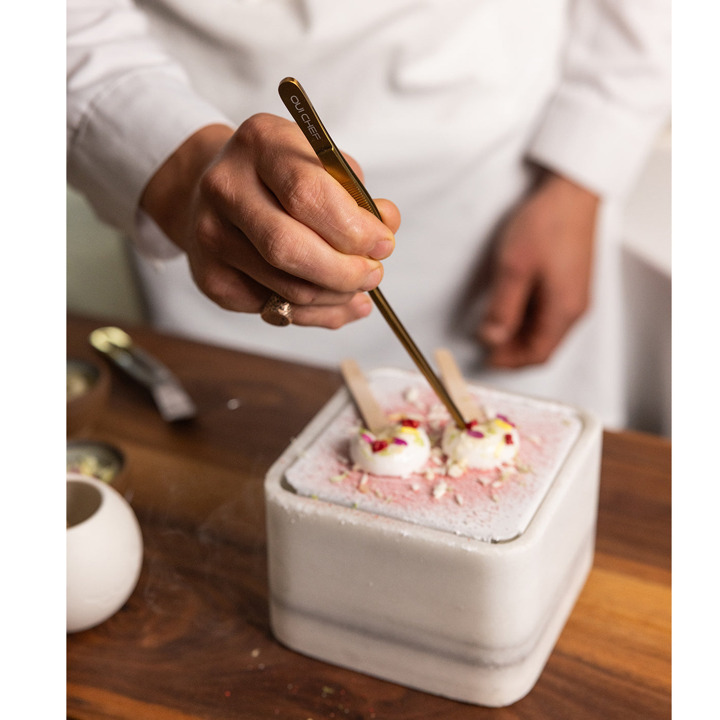 Person garnishing a dessert with a gold tweezers on a wooden table.