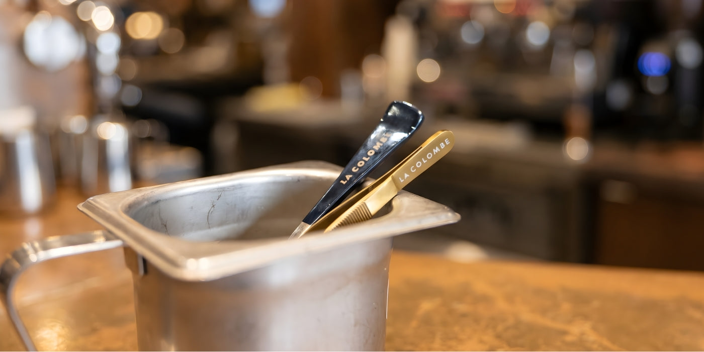 A Blue top chef's spoon and gold top plating tweezers standing in a stainless steel mise en place container in a professional kitchen.