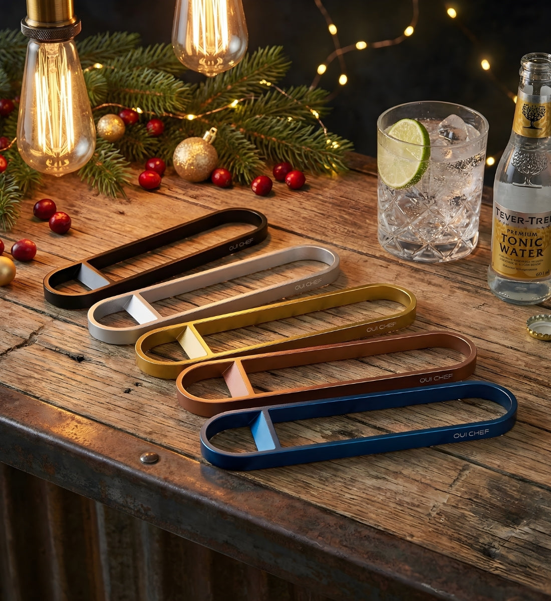 Set of five stainless steel Oui Chef bottle openers in different colors on a wooden surface with Christmas decorations and a gin and tonic drink.