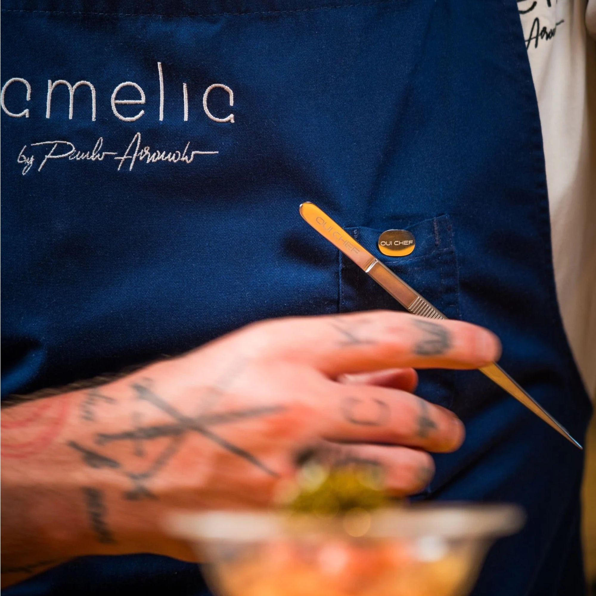 Chef wearing a blue apron with 'Amelia' branding attaching  small stainless steel tweezers to the holdfast magnetic clip