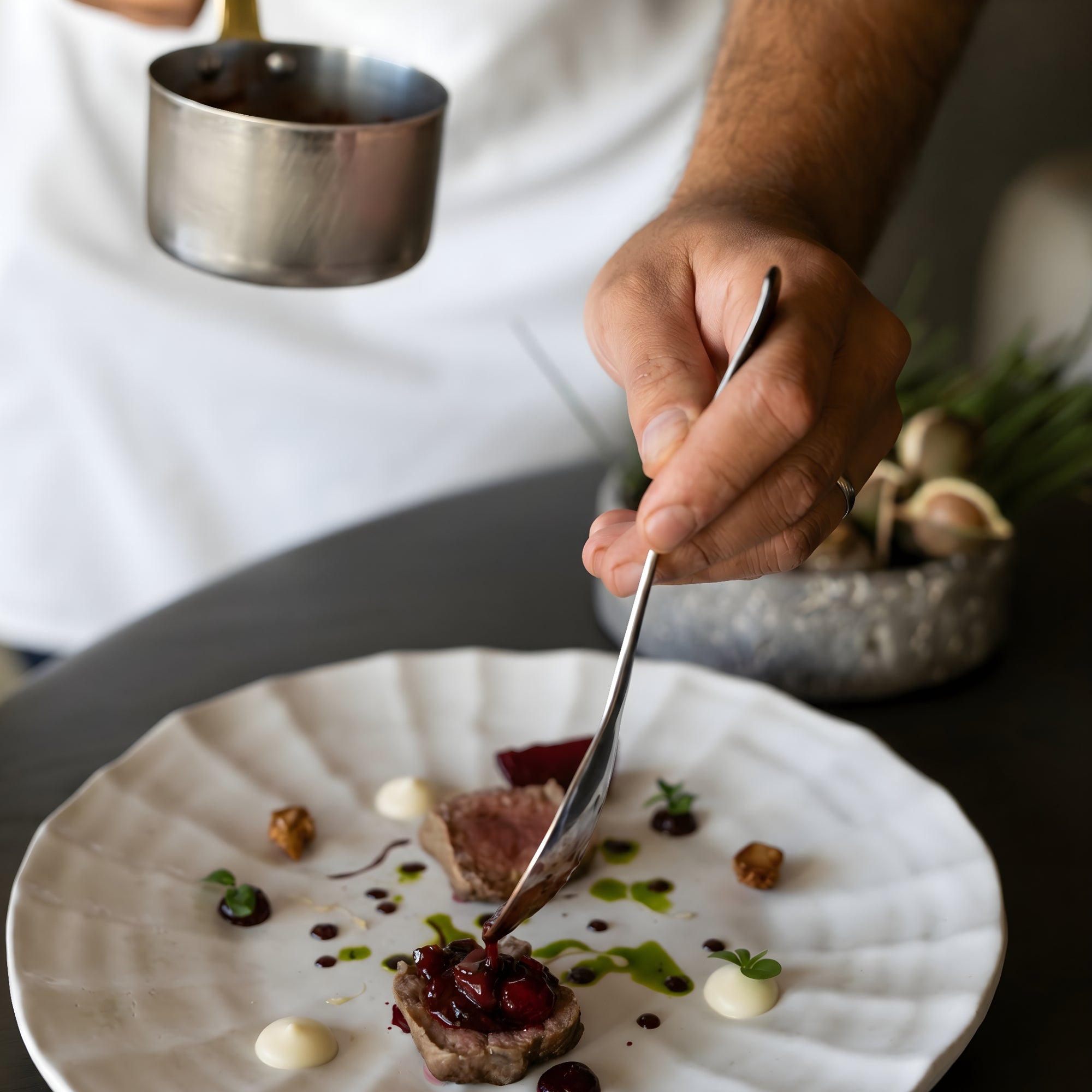 Chef using a spoon to coat a plated piece of meat with a textured gourmet sauce