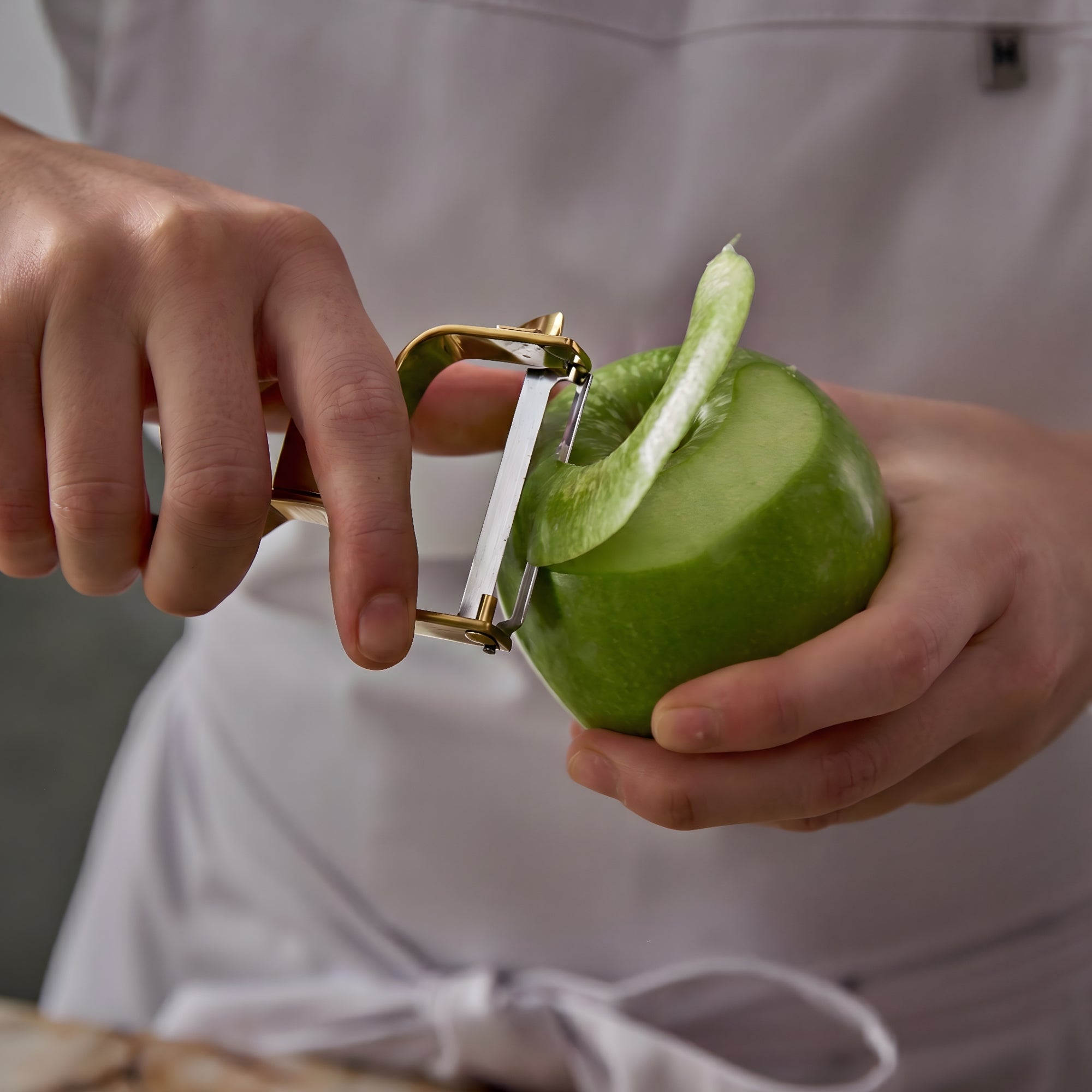 Chef carefully peeling a fresh apple with a gold stainless steel peeler in a modern kitchen with soft natural lighting and a premium culinary atmosphere.