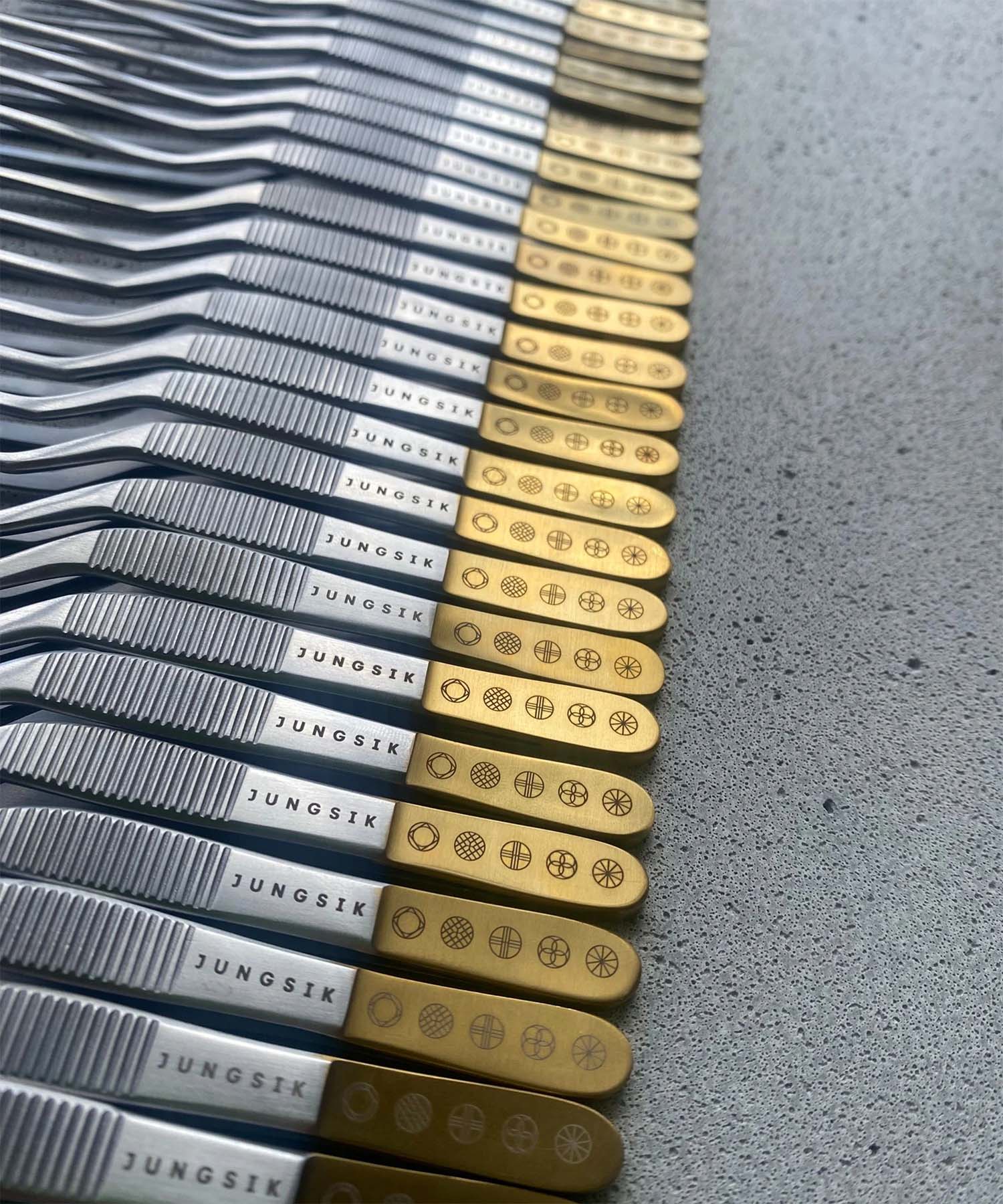 Thirty gold top offset tweezers sitting on a concrete slab with the Jungsik restaurant logo engraved.